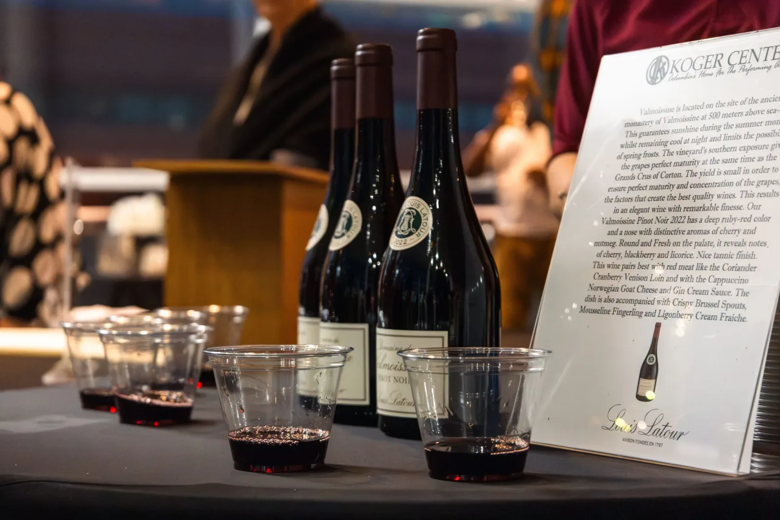 Three bottles of wine and 5 cups of wine on a table with a black tablecloth at a wine tasting.
