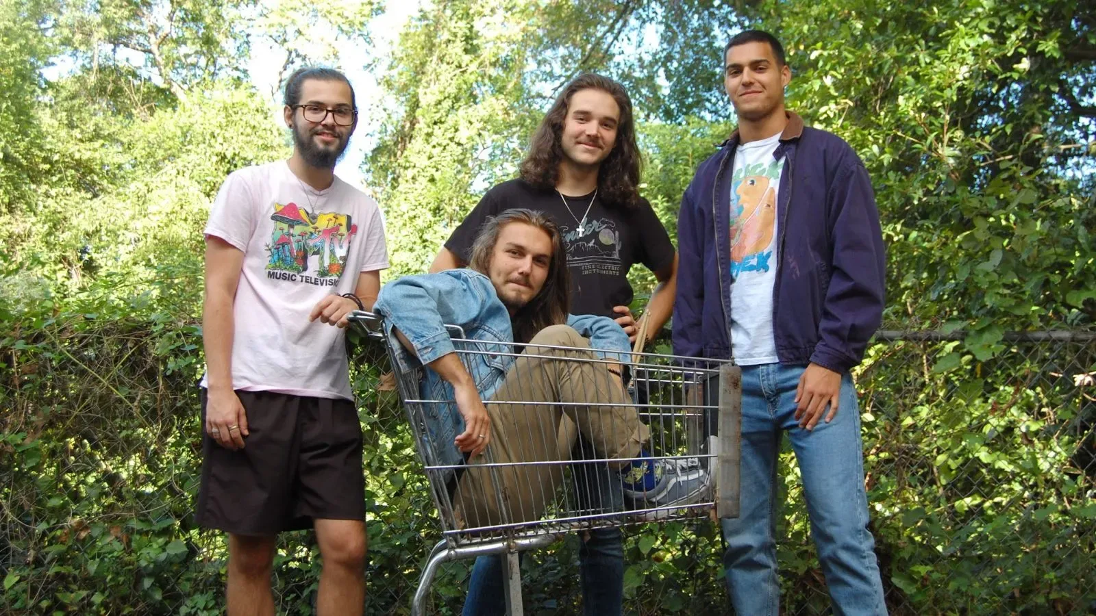 A group of four men; one is sitting in a grocery cart, three are standing behind the cart and against a forest background.
