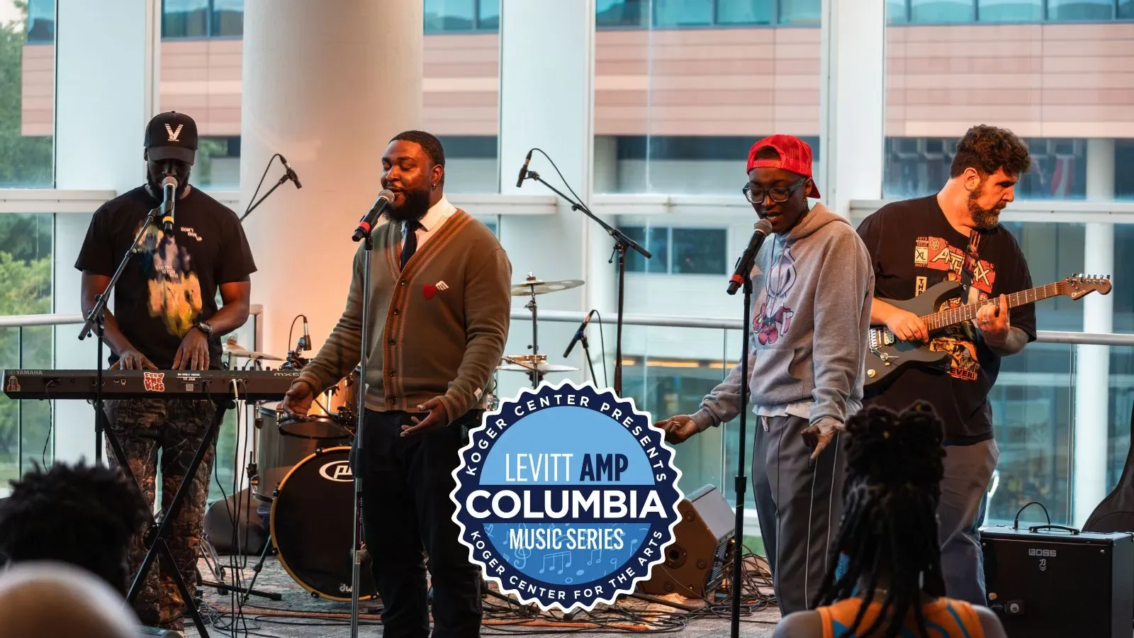 A group of four musicians performs on an indoor stage with large windows behind them. One person sings into a microphone at center while others play keyboard, guitar, and bass. A “Levit Amp Columbia Music Series” logo is displayed in front of the stage.