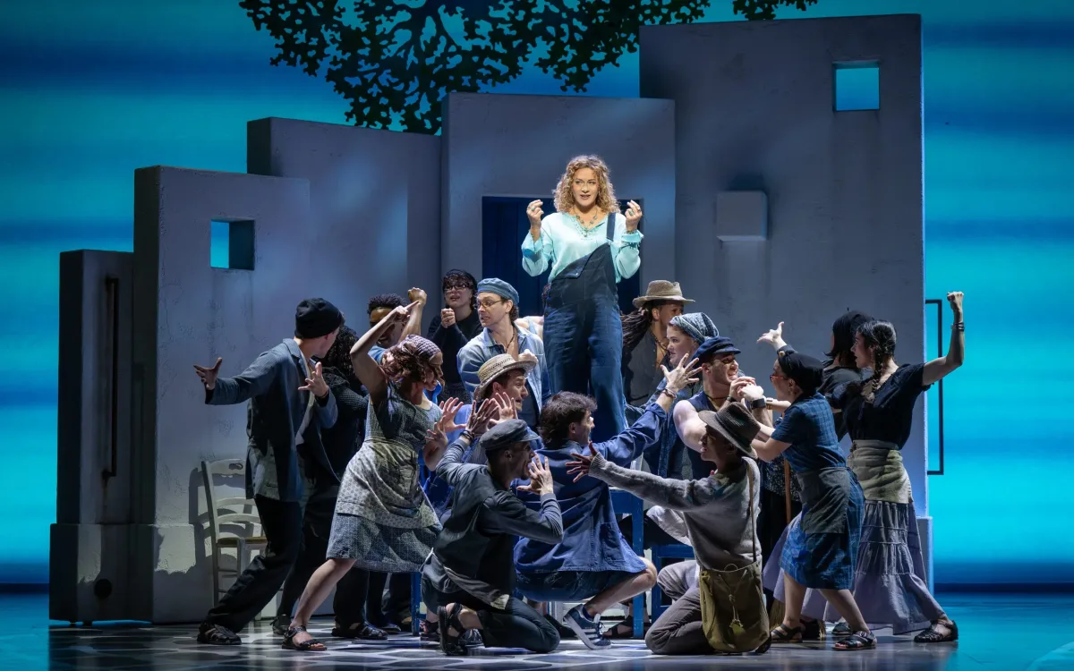 A group of dancers surrounding a woman in overalls and a blue shirt standing on a table.