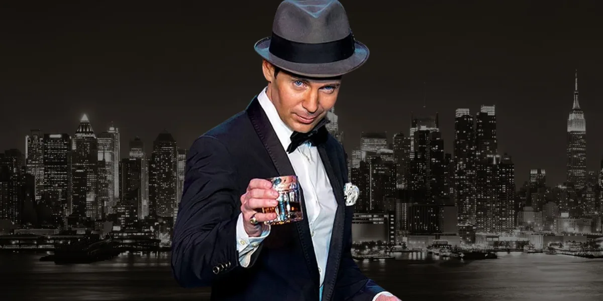 A photo of a man dressed like Frank Sinatra holding a rocks glass in front of a black and white city skyline.