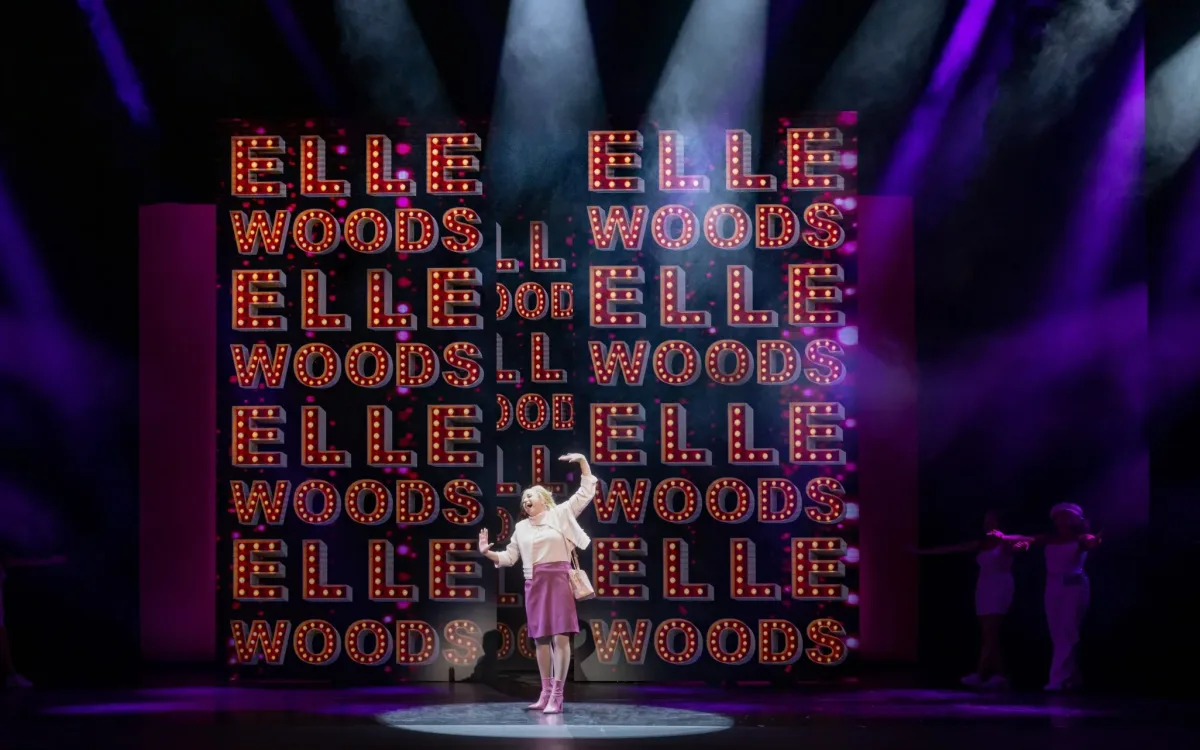 A blonde woman standing and singing in a spotlight. The walls behind her are covered with 