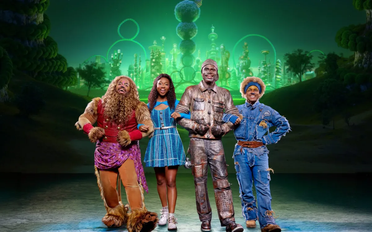 A group of actors dressed like characters from the Wizard of Oz walking with arms interlocked in front of a green background.
